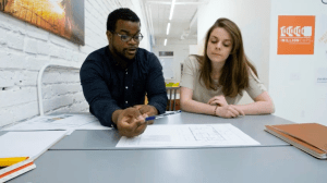 Two people sit at a table. The left person has a pencil in hand. The right is listening.