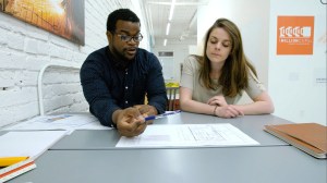 Two people sit at a table. The left person has a pencil in hand. The right is listening.