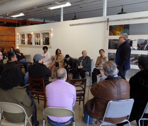 A person stands to ask a panel of 5 entrepreneurs a question. Other adults sit and listen around them.