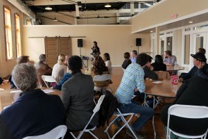 A facilitator stands at the front of a room of people sitting at circular tables